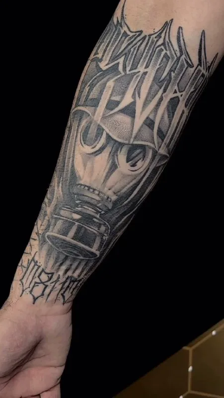 Máscara y letras en el antebrazo Tatuaje de una máscara y letras estilizadas en negro y gris en el antebrazo.